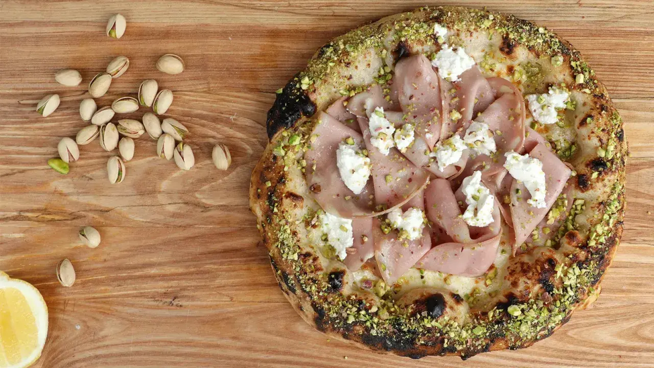 Pizza mit Mortadella, Pistazien und cremigem Käse, daneben einige Pistazien und eine Zitronenspalte.