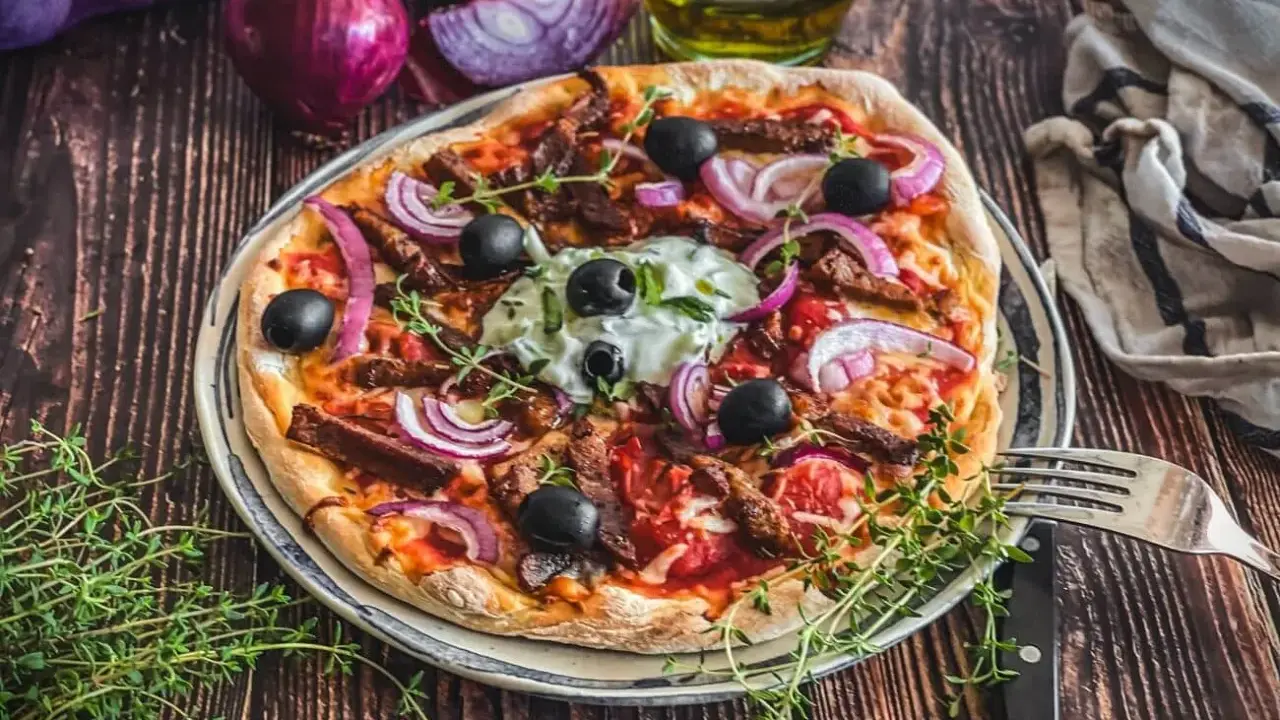Herzhafte Gyros Pizza mit Zwiebeln, Oliven und Kräutern, serviert auf einem Teller mit Gabel und Messer.