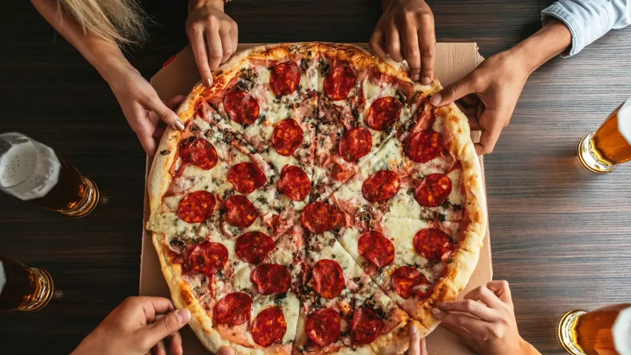 Hände greifen nach einer großen Salamipizza, die auf einem Tisch liegt. Jeder bekommt seinen fairen Anteil an pizzateig pro person.