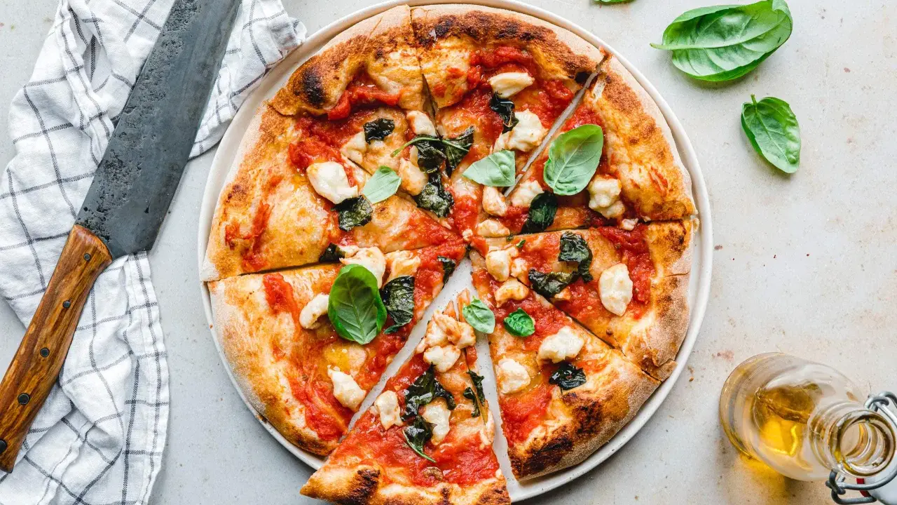 Eine köstliche, frisch gebackene pizza vegana mit Tomaten, Basilikum und veganem Mozzarella. Daneben ein Messer und ein Glas Öl.