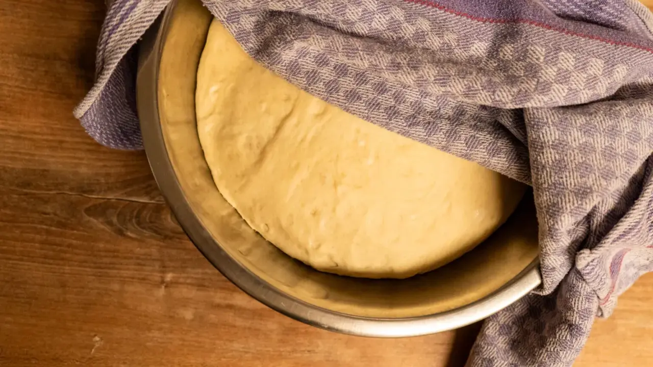 Frischer, aufgegangener Pizzateig ruht in einer Schüssel, bedeckt mit einem gemusterten Tuch. Perfekt für Ihr Pizzateig Rezept.