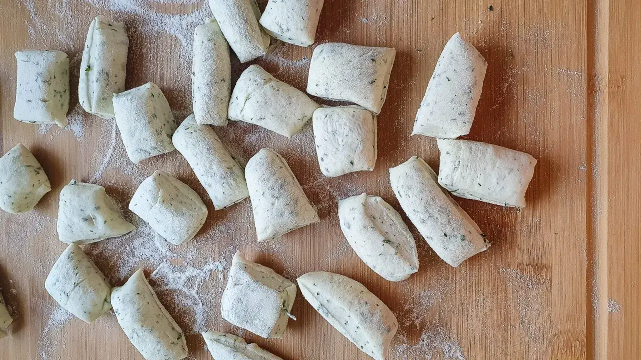 Vorbereitung für ein Grissini Rezept: Viele kleine, unregelmäßig geformte Teiglinge mit Kräutern liegen auf einem Holzbrett, bestäubt mit Mehl.