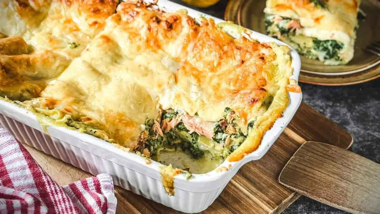 Herzhafte Lachs Spinat Lasagne, goldbraun überbacken, dampft in einer weißen Auflaufform. Ein Stück auf einem Teller daneben.