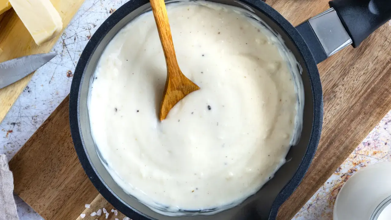 Cremige Bechamelsauce in einer Pfanne, bereit für die Lasagne. Ein Holzlöffel rührt die sämige Masse.
