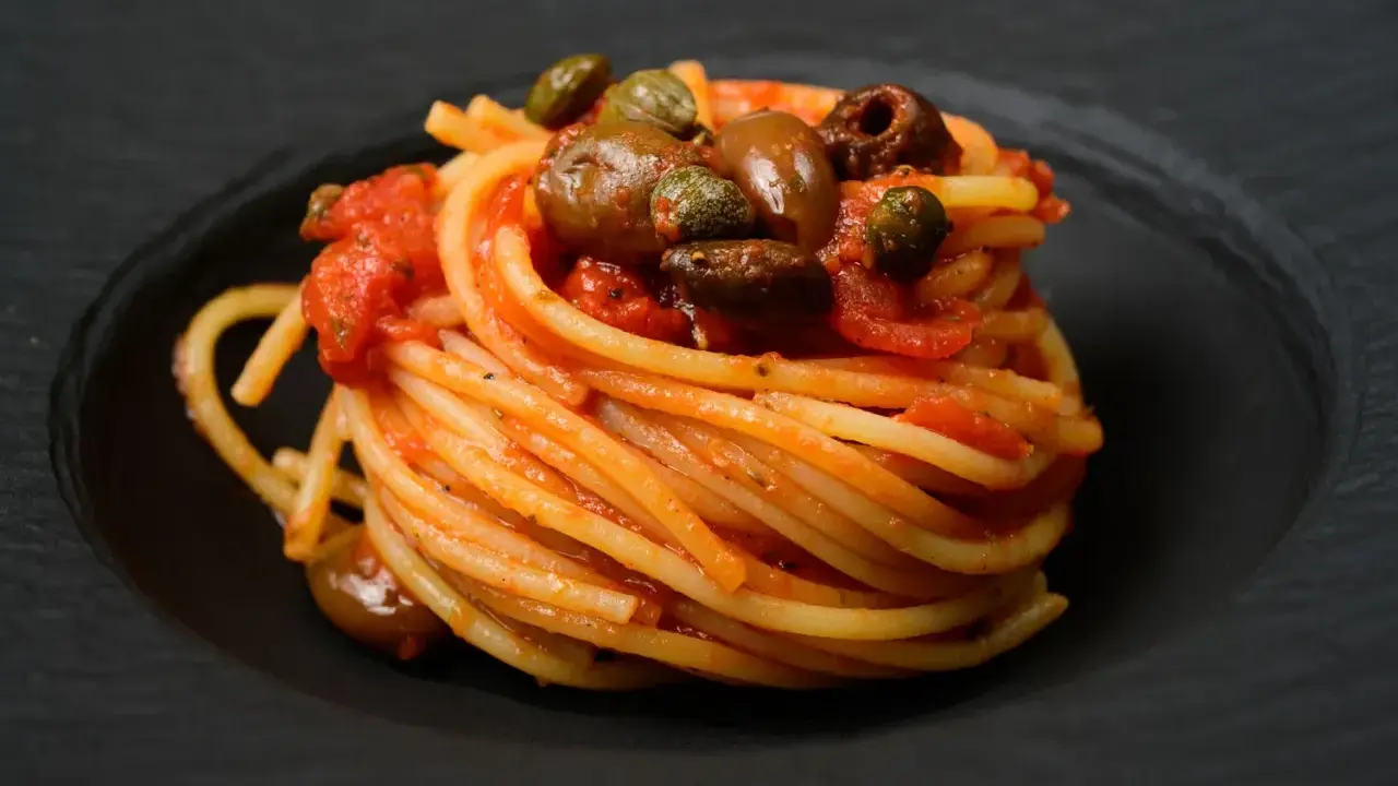 Ein kunstvoll aufgetürmter Teller Spaghetti Puttanesca, mit Oliven und Kapern garniert, auf schwarzem Teller.