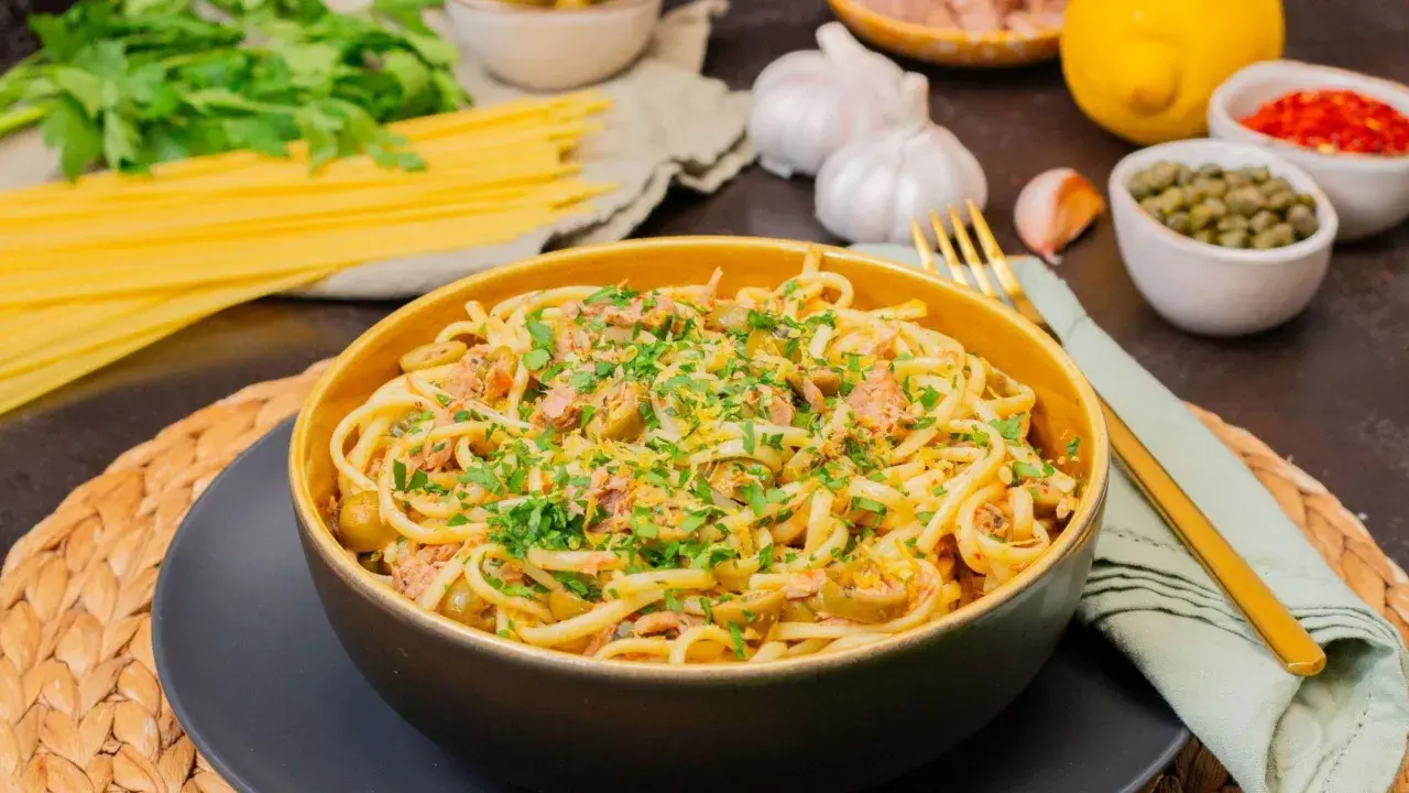 Eine Schüssel mit köstlicher Thunfisch Pasta, garniert mit Petersilie und Zitronenschale. Daneben liegen Spaghetti, Knoblauch und Kapern.