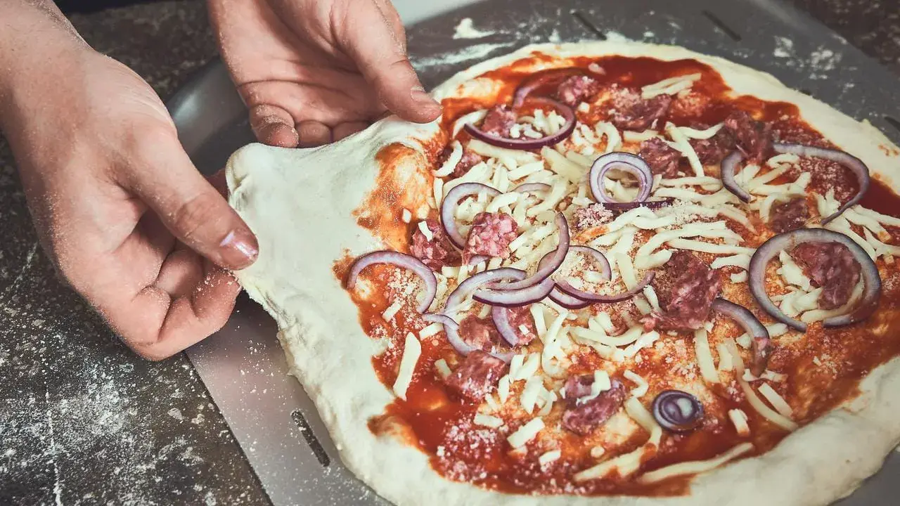 Hände schieben einen rohen Pizzaboden mit Tomatensauce, Käse, Salami und Zwiebeln auf einen Pizzaschieber. Der pizzateig klebt am schieber.
