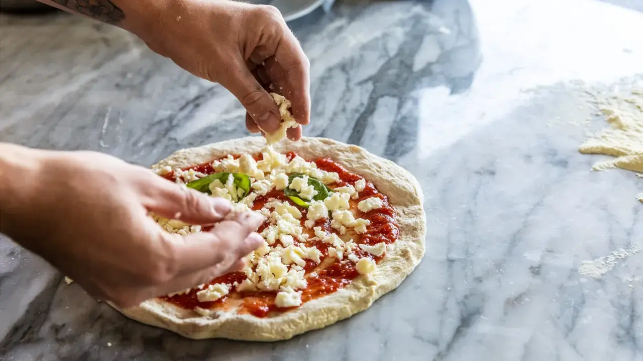 Hände belegen Pizza mit zerbröckeltem Käse, einer köstlichen fior di latte Alternative, auf Marmortisch.