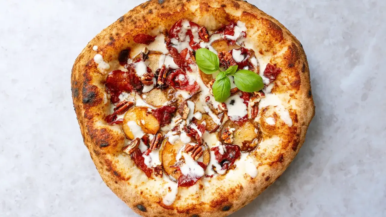 Pizza mit karamellisierten Zwiebeln, Speck, Pekannüssen und einem Hauch von Parmesan. Ein grünes Basilikumblatt krönt das Kunstwerk.