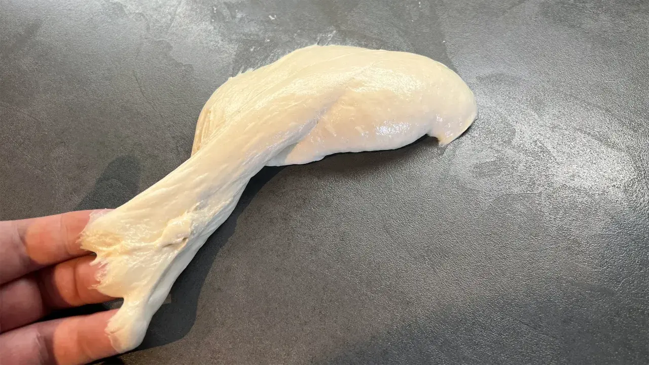 Ein Stück heller, klebriger Pizzateig wird von einer Hand gehalten und dehnt sich auf einer dunklen Arbeitsfläche aus.