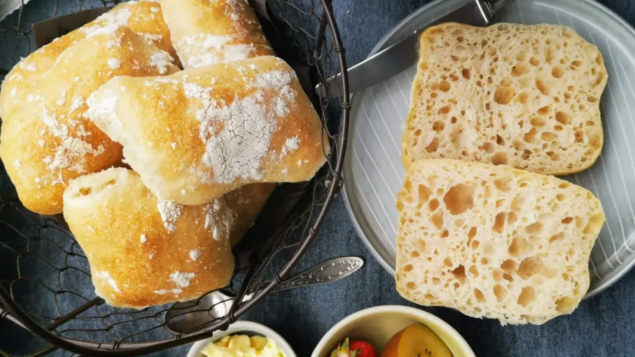 Frisch gebackene Ciabatta-Brötchen, bestäubt mit Mehl, liegen in einem Korb. Daneben zwei aufgeschnittene Scheiben mit luftiger Krume – perfekt zum ciabatta selber backen.