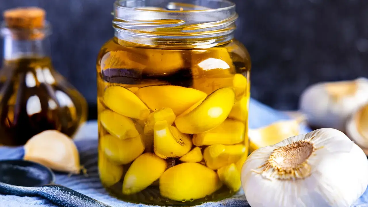 Ein Glas mit eingelegtem Knoblauch, typisch italienisch, gefüllt mit goldgelbem Öl. Daneben frische Knoblauchzehen und eine Ölflasche.