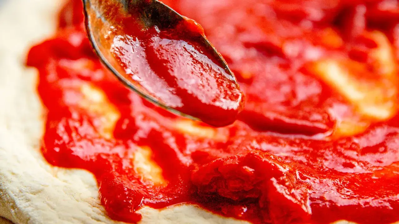 Eine Löffelspitze verteilt die leuchtend rote Tomatensauce auf dem Teig für eine authentische Pizza Napoletana.