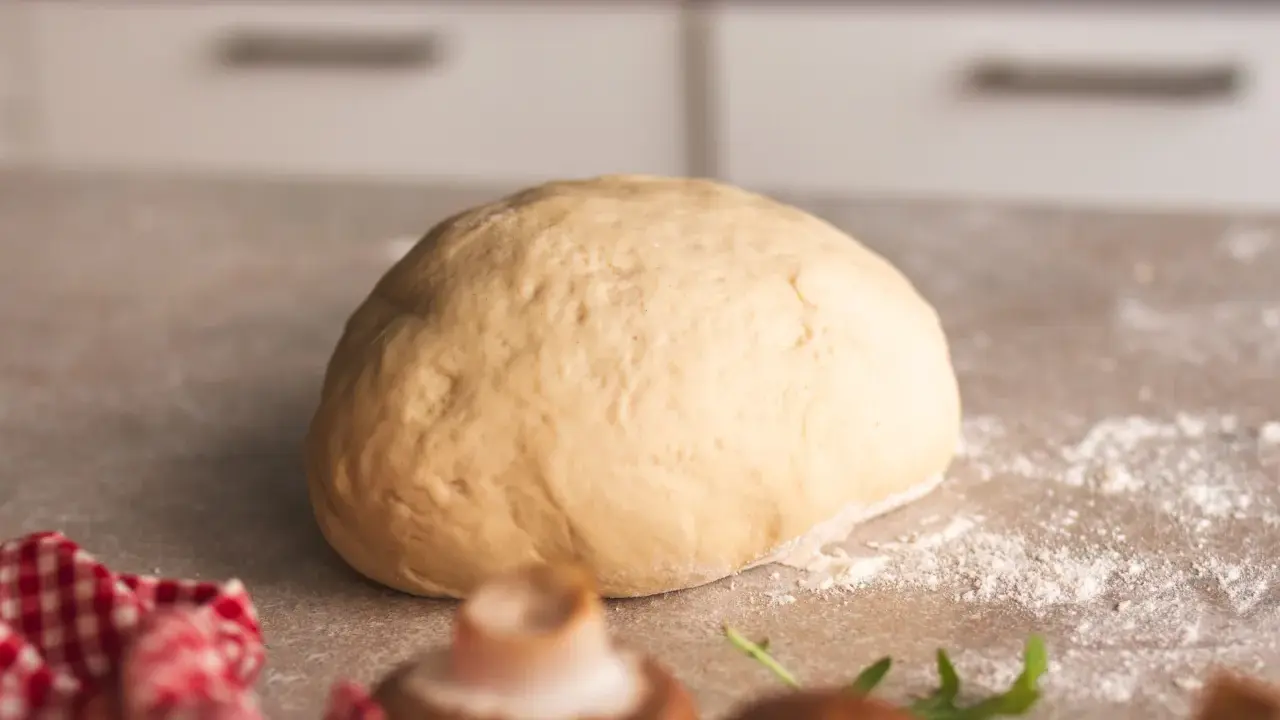 Frischer, goldgelber Pizzateig liegt bereit zum Formen. Ein paar Pilze und Rucola deuten auf eine leckere Pizza hin.