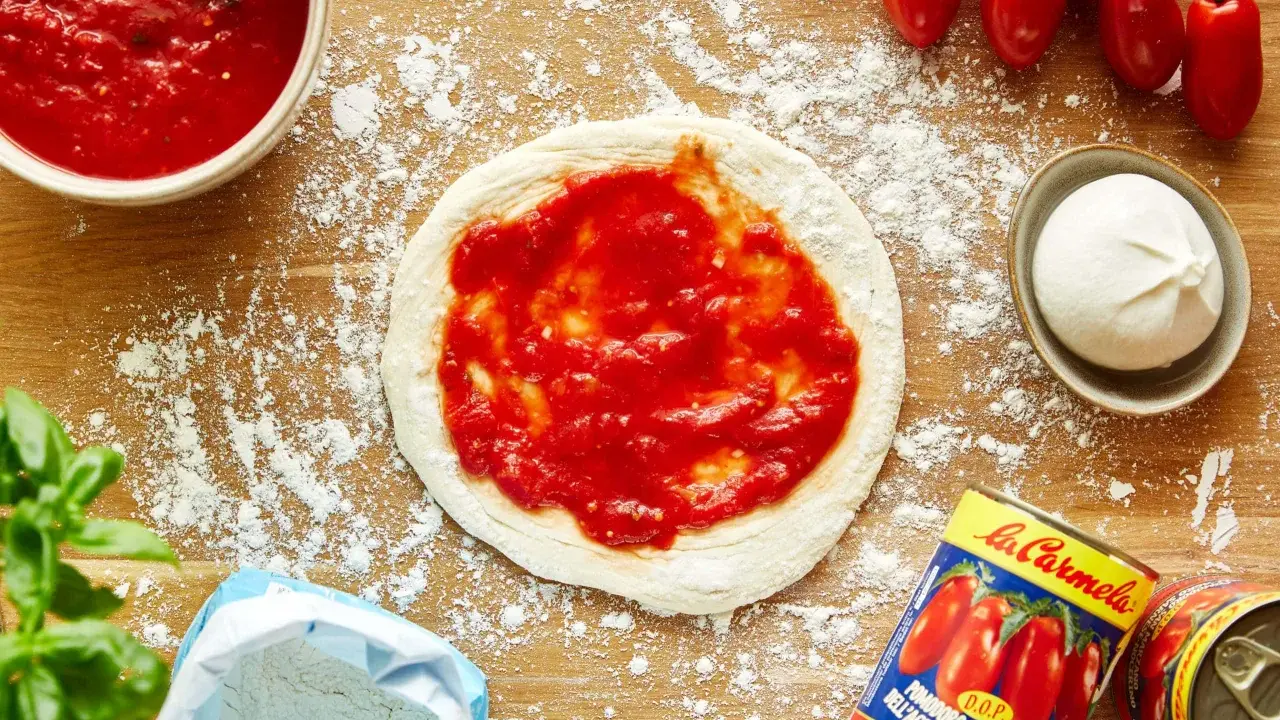 Pizza-Zutaten auf Holzbrett: Teig mit Tomatensauce, Mozzarella, frische Tomaten, Basilikum, Mehl und eine Dose San Marzano Tomaten. Die Frage "wieviel Tomatensauce pro Person" wird hier visuell beantwortet.