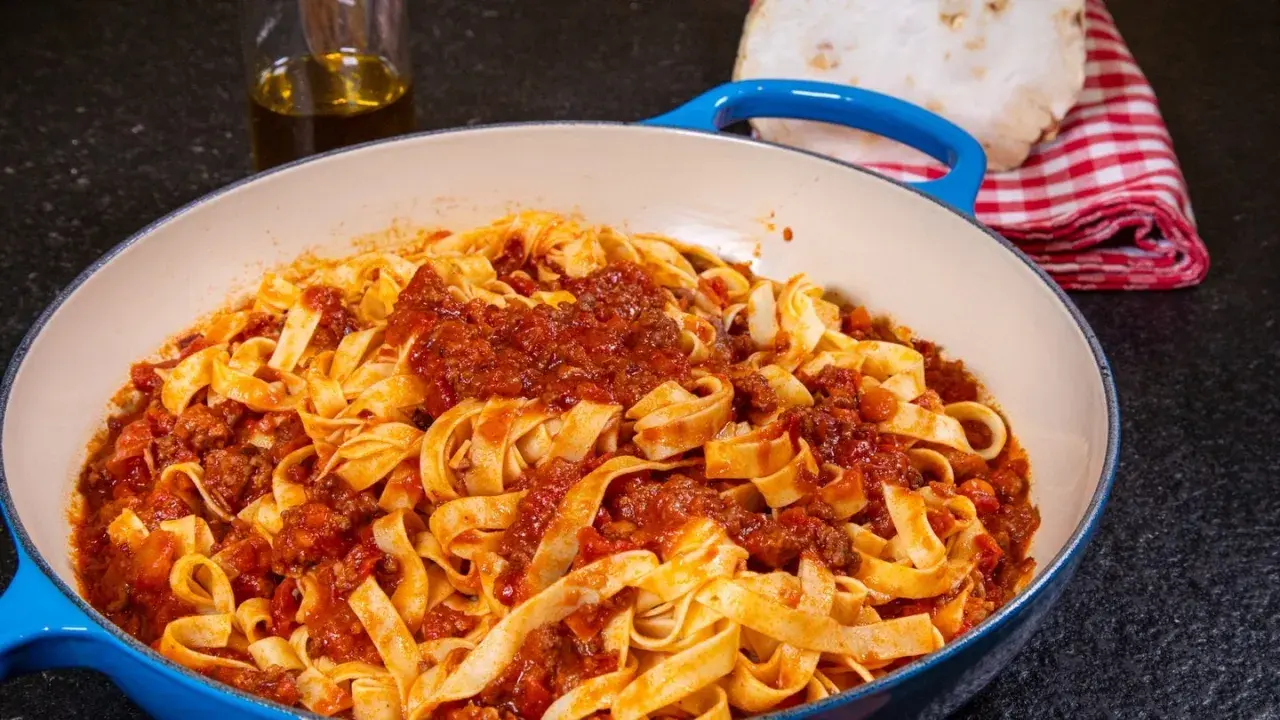 Herzhaftes Tagliatelle Bolognese Rezept in einer blauen Pfanne, mit Olivenöl und einem karierten Tuch im Hintergrund.