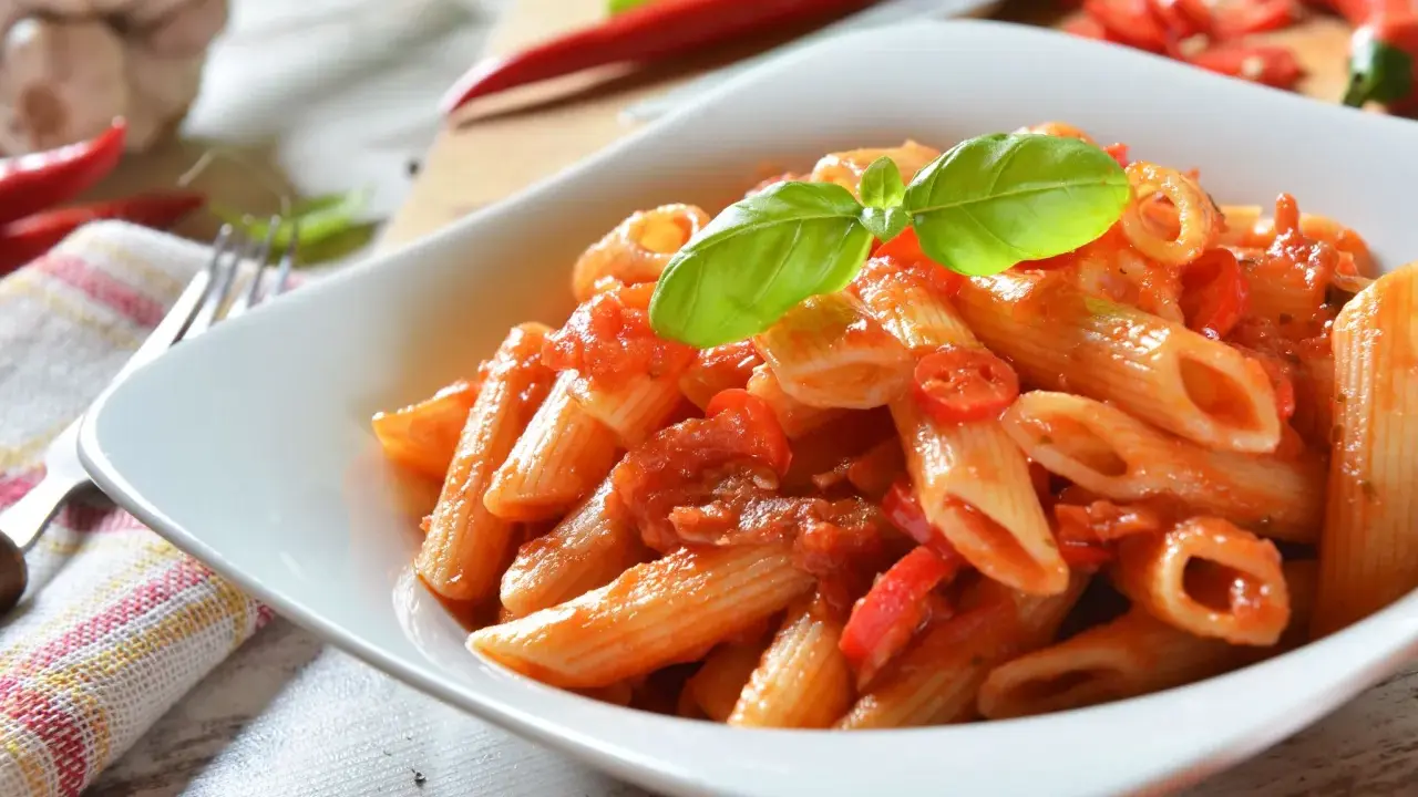 Eine Schüssel Penne Arrabiata, garniert mit Basilikum, mit scharfen Chilischoten und Knoblauch im Hintergrund.