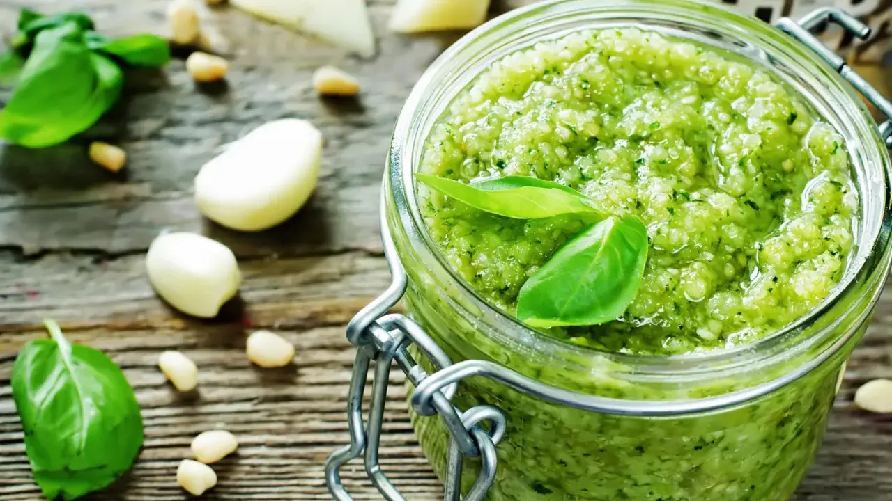 Frisches Pesto-Rezept im Glas, umgeben von Basilikum, Pinienkernen, Knoblauch und Parmesan auf rustikalem Holz.