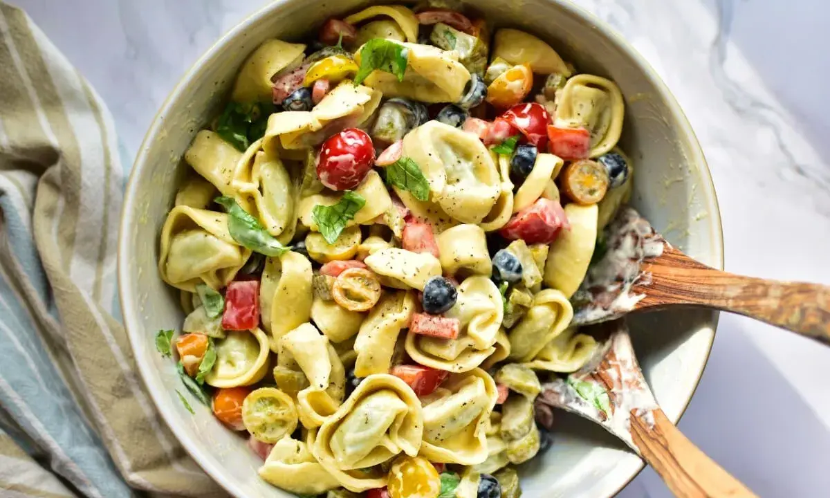 Ein cremiger Tortellini Salat mit Kirschtomaten, Oliven und Kräutern, serviert in einer Schüssel mit Holzspateln.