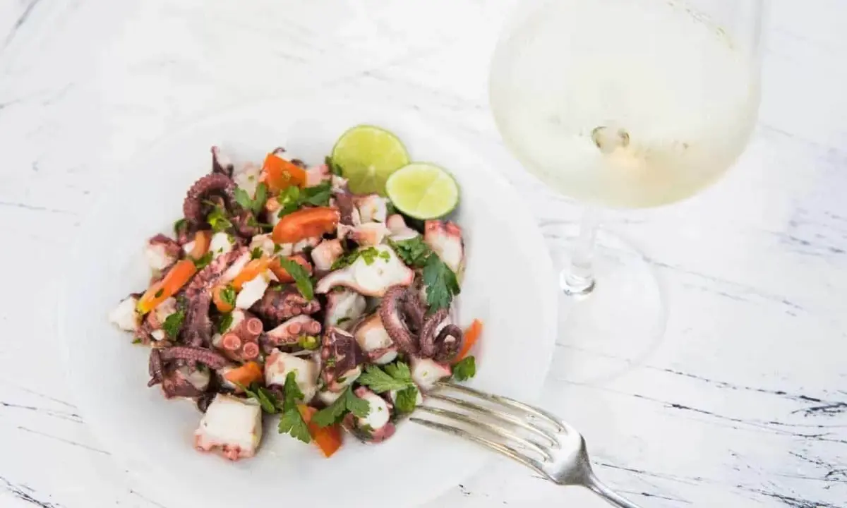Frischer Insalata di polpo mit Tomaten, Petersilie und Limettenspalten auf einem Teller, daneben ein Glas Weißwein.