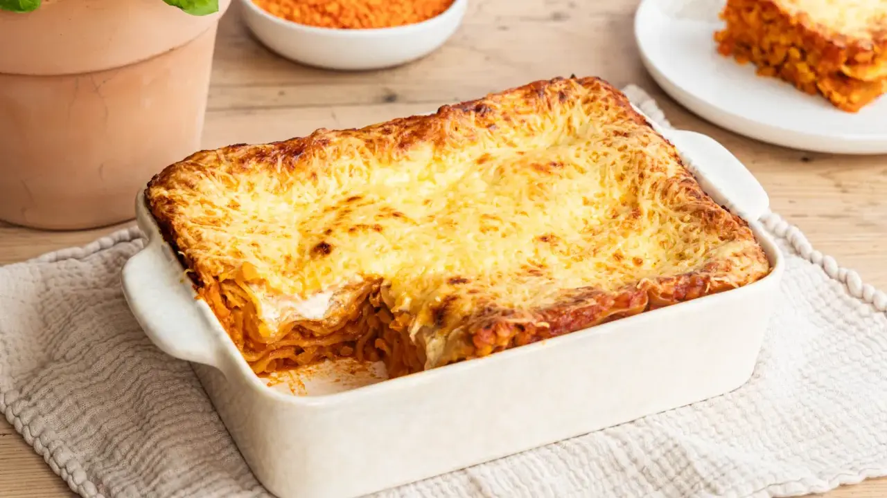 Herzhafte Linsen Lasagne mit goldbraun überbackener Käseschicht, daneben eine Schüssel roter Linsen und ein Teller mit einem Stück Lasagne.