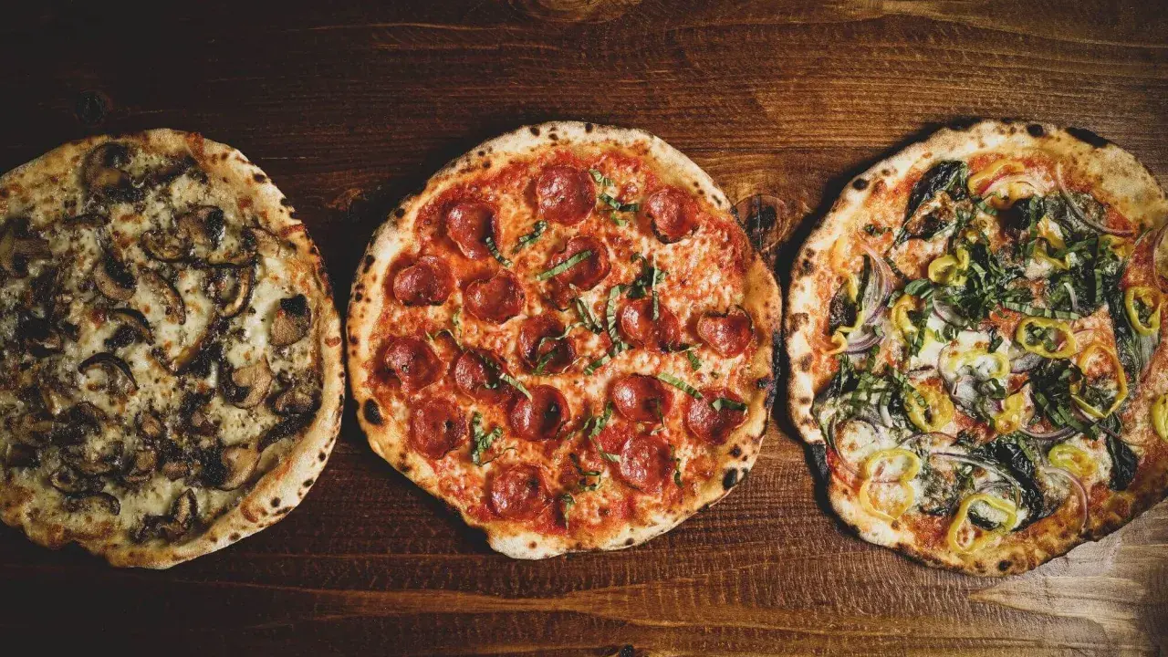 Drei leckere Pizza-Sorten: eine mit Pilzen, eine mit Salami und eine mit Gemüse, liegen nebeneinander auf einem Holztisch.