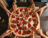 Hände greifen nach einer großen Salamipizza, die auf einem Tisch liegt. Jeder bekommt seinen fairen Anteil an pizzateig pro person.