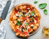 Eine köstliche, frisch gebackene pizza vegana mit Tomaten, Basilikum und veganem Mozzarella. Daneben ein Messer und ein Glas Öl.