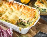 Herzhafte Lachs Spinat Lasagne, goldbraun überbacken, dampft in einer weißen Auflaufform. Ein Stück auf einem Teller daneben.