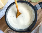 Cremige Bechamelsauce in einer Pfanne, bereit für die Lasagne. Ein Holzlöffel rührt die sämige Masse.