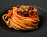Ein kunstvoll aufgetürmter Teller Spaghetti Puttanesca, mit Oliven und Kapern garniert, auf schwarzem Teller.