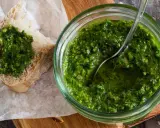 Frisches schnittlauch pesto in einem Glas, mit einem Löffel darin. Ein Stück Brot mit dem grünen Pesto darauf liegt daneben.