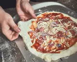 Hände schieben einen rohen Pizzaboden mit Tomatensauce, Käse, Salami und Zwiebeln auf einen Pizzaschieber. Der pizzateig klebt am schieber.