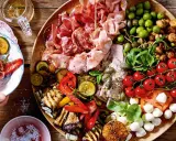 Eine üppige italienische Antipasti-Platte wird liebevoll dekoriert: Wurst, Käse, Oliven, gegrilltes Gemüse und frische Kräuter laden zum Genuss ein.
