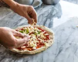 Hände belegen Pizza mit zerbröckeltem Käse, einer köstlichen fior di latte Alternative, auf Marmortisch.