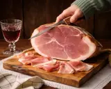 Ein Messer schneidet hauchdünne Scheiben von einem ganzen Schinken, **prosciutto crudo**, auf einem Holzbrett. Daneben ein Glas Rotwein.