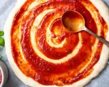 Eine Pizza nur mit Tomatensauce wird mit einer Löffelspirale belegt. Frisches Basilikum liegt daneben.