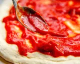 Eine Löffelspitze verteilt die leuchtend rote Tomatensauce auf dem Teig für eine authentische Pizza Napoletana.