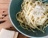 Ein Teller cremiges Cacio e Pepe, bestreut mit frisch geriebenem Pecorino und schwarzem Pfeffer. Daneben liegen Pfefferkörner und ein Stück Pecorino.
