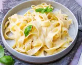 Cremige Pasta Alfredo mit frischem Basilikum und schwarzem Pfeffer, serviert auf einem grauen Tischtuch.