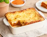 Herzhafte Linsen Lasagne mit goldbraun überbackener Käseschicht, daneben eine Schüssel roter Linsen und ein Teller mit einem Stück Lasagne.