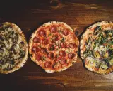 Drei leckere Pizza-Sorten: eine mit Pilzen, eine mit Salami und eine mit Gemüse, liegen nebeneinander auf einem Holztisch.