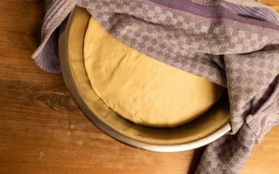 Frischer, aufgegangener Pizzateig ruht in einer Schüssel, bedeckt mit einem gemusterten Tuch. Perfekt für Ihr Pizzateig Rezept.