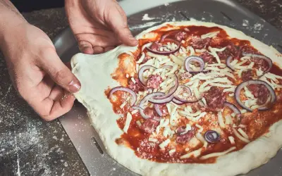 Hände schieben einen rohen Pizzaboden mit Tomatensauce, Käse, Salami und Zwiebeln auf einen Pizzaschieber. Der pizzateig klebt am schieber.
