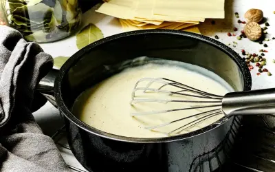 Cremige Béchamelsauce wird in einem Topf mit einem Schneebesen zubereitet. Daneben liegen Lasagneplatten und Gewürze.
