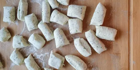 Vorbereitung für ein Grissini Rezept: Viele kleine, unregelmäßig geformte Teiglinge mit Kräutern liegen auf einem Holzbrett, bestäubt mit Mehl.