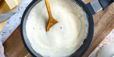 Cremige Bechamelsauce in einer Pfanne, bereit für die Lasagne. Ein Holzlöffel rührt die sämige Masse.