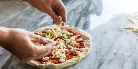 Hände belegen Pizza mit zerbröckeltem Käse, einer köstlichen fior di latte Alternative, auf Marmortisch.