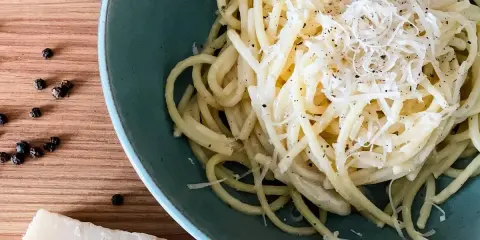 Ein Teller cremiges Cacio e Pepe, bestreut mit frisch geriebenem Pecorino und schwarzem Pfeffer. Daneben liegen Pfefferkörner und ein Stück Pecorino.