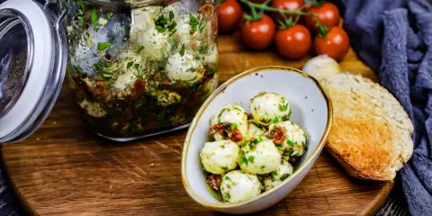 Marinierte Mozzarellakugeln in einem Glas, bereit für köstliche Mozzarella Rezepte. Dazu gibt es frische Tomaten und geröstetes Brot.